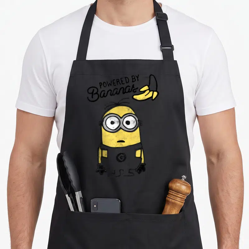 Despicable jook apron - Minion character below arched POWERED BY Bananas text and yellow banana peel — shown on a black cotton canvas apron against a white background