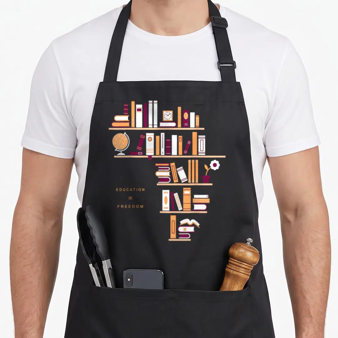 Africa education apron - Minimalist bookshelf with books, globe, and text EDUCATION IS FREEDOM in orange and purple — shown on a black cotton canvas apron against a white background