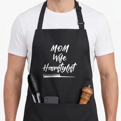 Hairstylist mother's day aprons - White script text MOM Wife Hairstylist with a horizontal comb illustration below — shown on a black cotton canvas apron against a white background