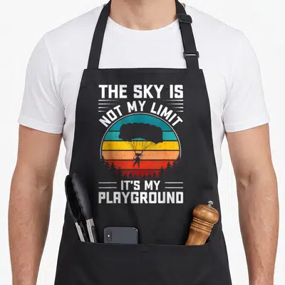 Limit playground sky apron - Retro sun graphic with parachutist silhouette and white text over teal orange and red — shown on a black cotton canvas apron against a white background
