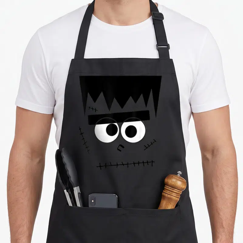 Munster halloween apron - Cartoon grey monster face with black jagged hair and stitched cheeks — shown on a black cotton canvas apron against a white background