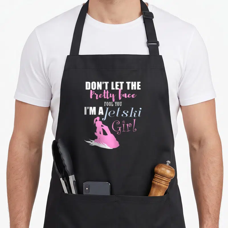 Jetski holiday apron - Pink jet ski girl silhouette with white and pink stacked typography and water splashes — shown on a black cotton canvas apron against a white background
