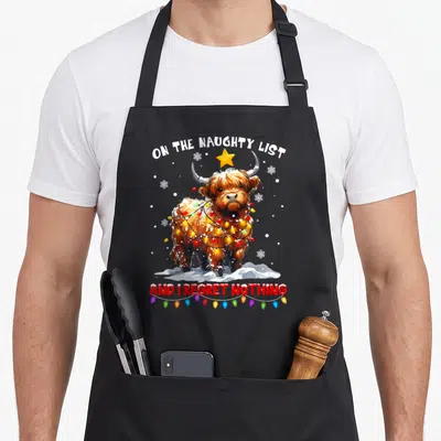 Naughty highland cow apron - Cartoon highland cow on snow with lights, star, and text ON THE NAUGHTY LIST AND I REGRET — shown on a black cotton canvas apron against a white background