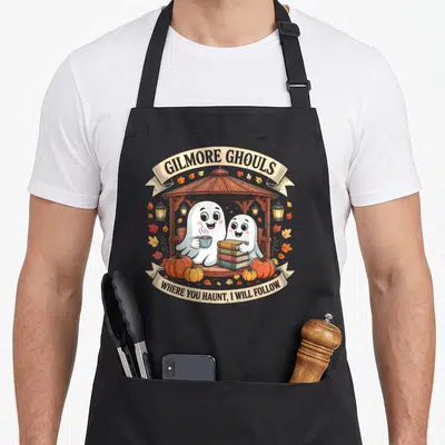 Gilmore halloween apron - Two white ghosts in a gazebo holding books and coffee with orange pumpkins and arched text — shown on a black cotton canvas apron against a white background