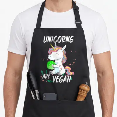 Herbivore veggie apron - Cartoon unicorn holding green cabbage with text unicorns are vegan and pink stars — shown on a black cotton canvas apron against a white background