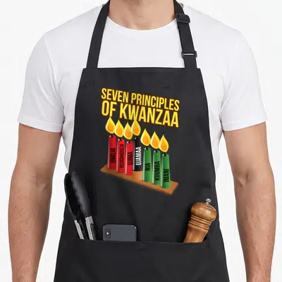 Seven funny kwanzaa apron - Seven Principles of Kwanzaa text above red black and green kinara candles with vertical — shown on a black cotton canvas apron against a white background