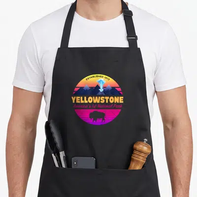National park 80s apron - Circular retro graphic of Yellowstone geyser, bison, and mountains over pink grid — shown on a black cotton canvas apron against a white background