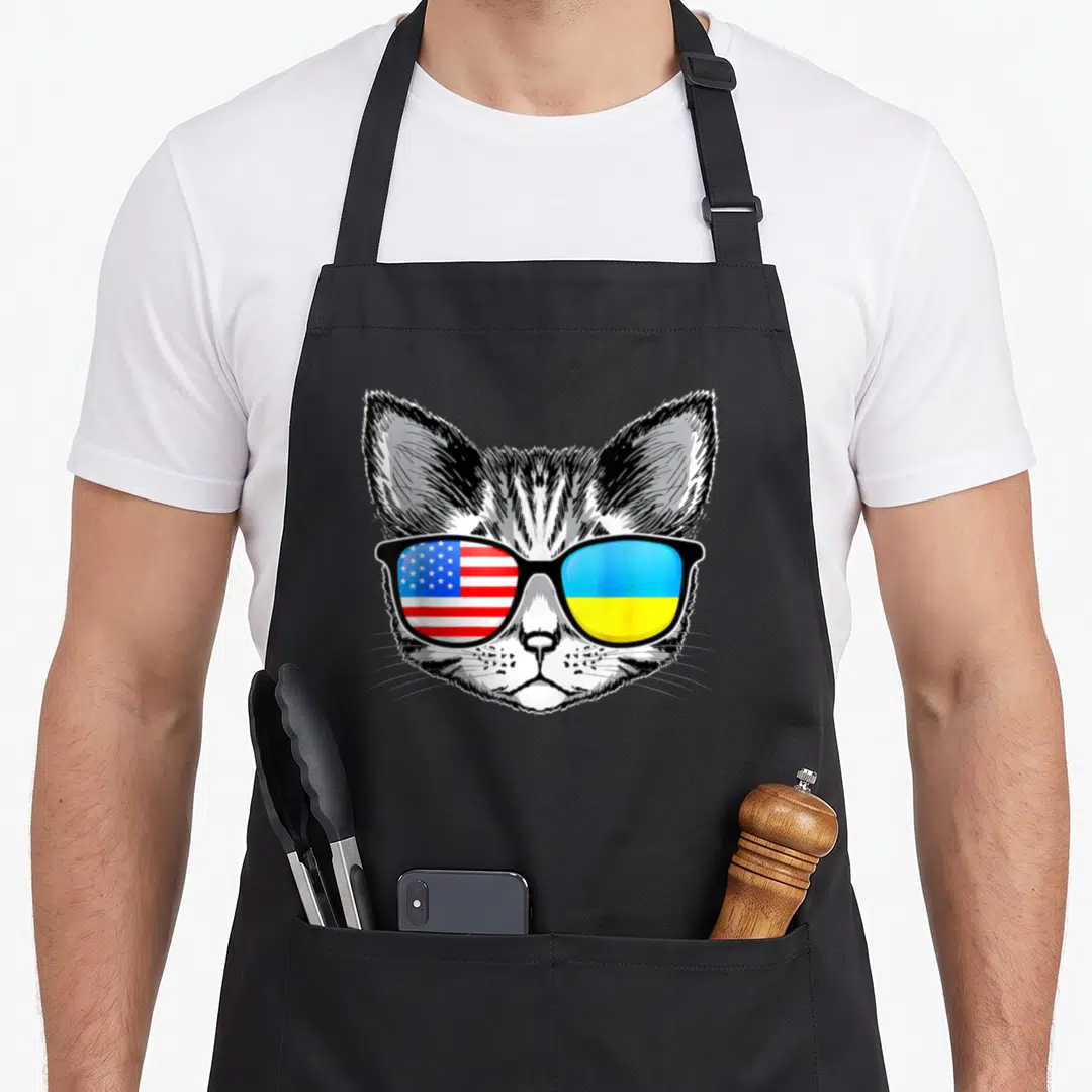 Ukraine root american flag apron - Cartoon cat head facing forward with sunglasses showing USA and Ukraine flags — shown on a black cotton canvas apron against a white background