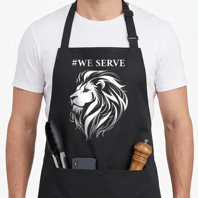 Lion to serve man apron - White line-art lion head profile with #WE SERVE text above — shown on a black cotton canvas apron against a white background