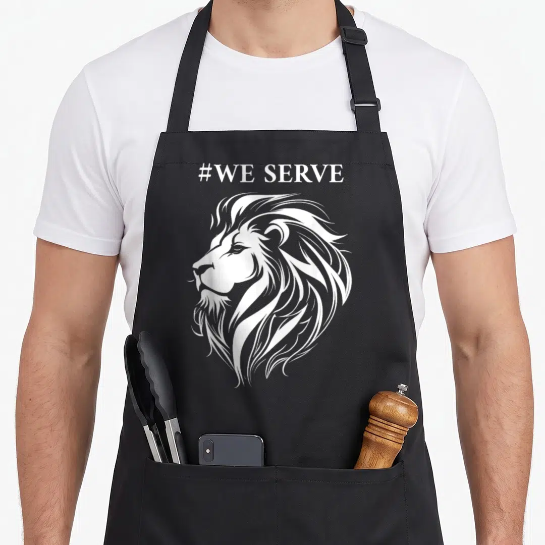Lion to serve man apron - White line-art lion head profile with #WE SERVE text above — shown on a black cotton canvas apron against a white background