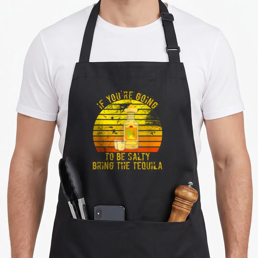 Salty sledding apron - Tequila bottle with sombrero and shot glasses against striped sunset with yellow text — shown on a black cotton canvas apron against a white background