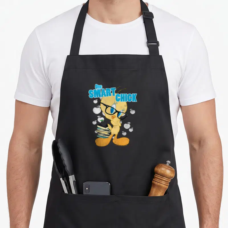 Chick smart apron - Tweety Bird wearing glasses holding books with apple on top and One SMART CHICK text — shown on a black cotton canvas apron against a white background