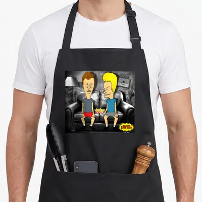 Butt judge humor apron - Beavis and Butt-Head sitting on black couch with chips, yellow logo bottom right — shown on a black cotton canvas apron against a white background