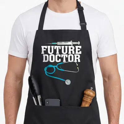 Training doctor apron - Future Doctor text with syringe and blue stethoscope on grey background — shown on a black cotton canvas apron against a white background
