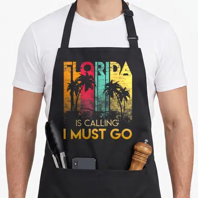 Florida summer aprons - Retro FLORIDA text in orange red teal yellow stripes with palm trees and IS CALLING I MUST GO — shown on a black cotton canvas apron against a white background