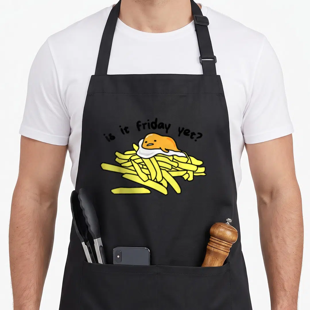 Friday egg apron - Cartoon fried egg on yellow fries with arched text is it Friday yet — shown on a black cotton canvas apron against a white background