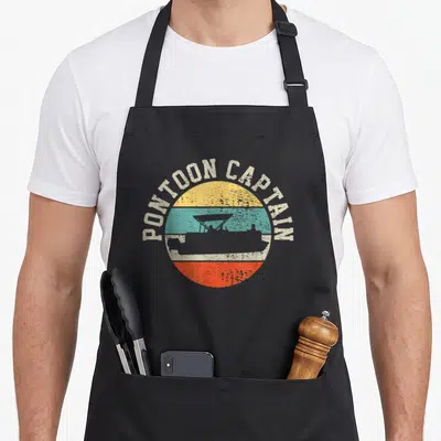 Captain boating boat apron - Retro PONTOON CAPTAIN text over yellow teal orange sunset circle with grey boat silhouette — shown on a black cotton canvas apron against a white background
