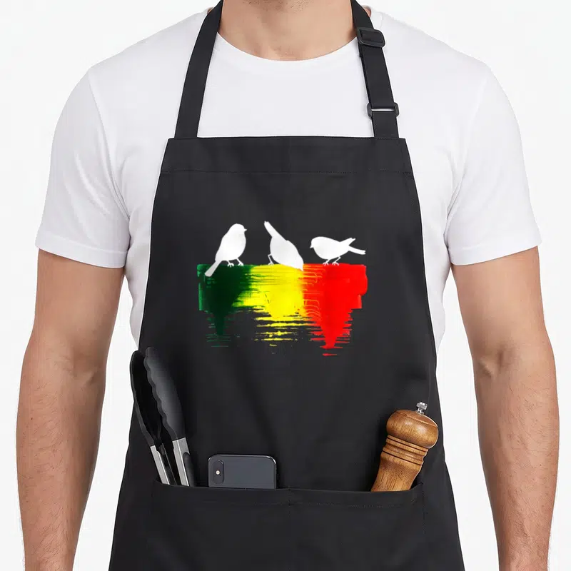Reggae bird apron - Three white bird silhouettes perched on a dripping green, yellow, and red paint stroke — shown on a black cotton canvas apron against a white background