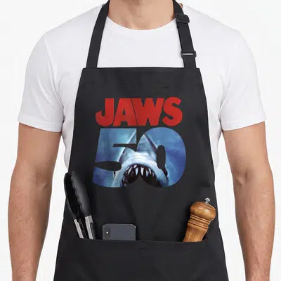 50th anniversary apron - Red JAWS text above blue 50 containing shark open mouth graphic — shown on a black cotton canvas apron against a white background