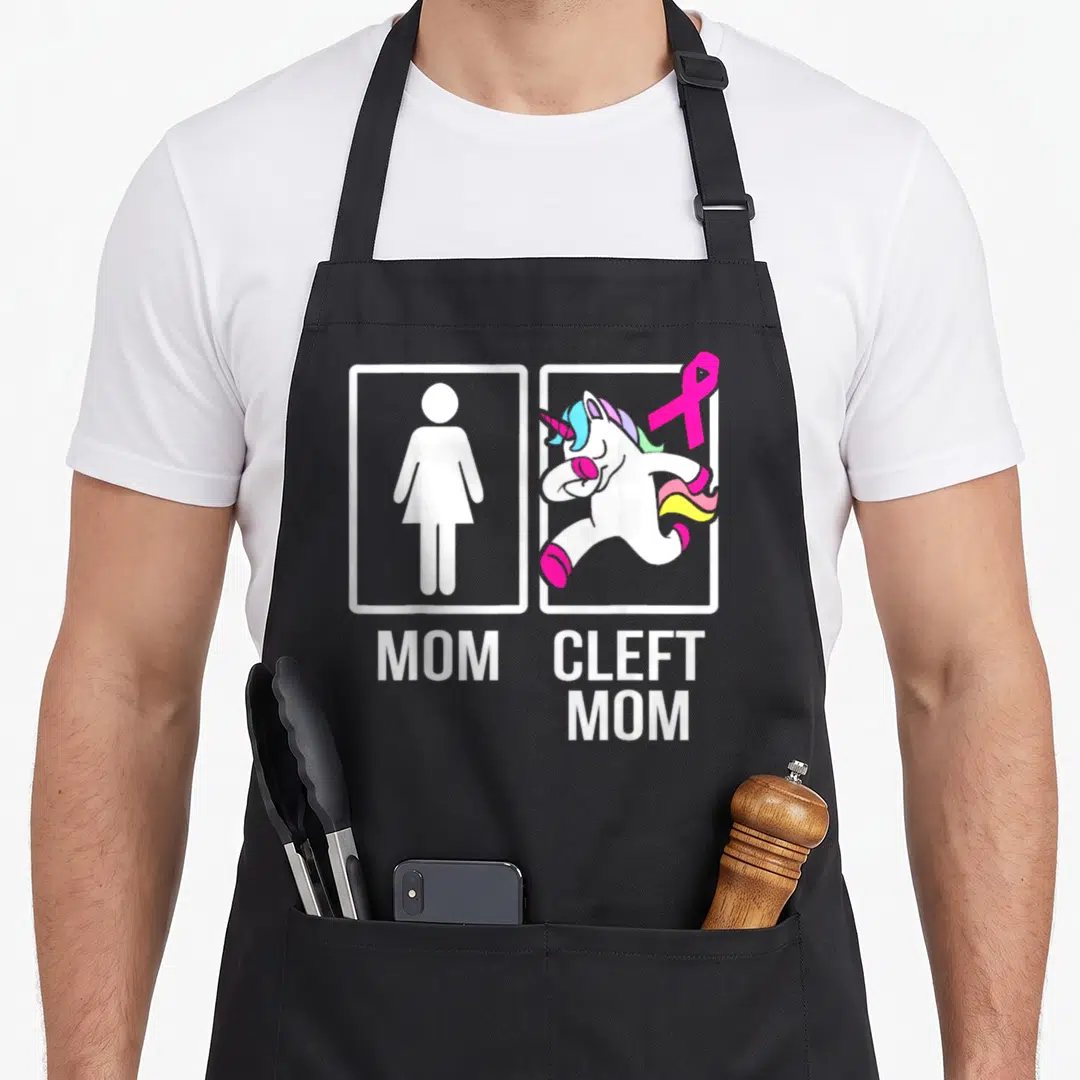 Cleft unicorn apron - Two white frames showing woman symbol and cartoon unicorn with pink ribbon and text MOM CLEFT MOM — shown on a black cotton canvas apron against a white background