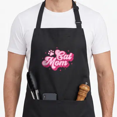 Mam kitty cat apron - Cat Mom text in light pink script on dark pink glittery shape with paw print and stars — shown on a black cotton canvas apron against a white background