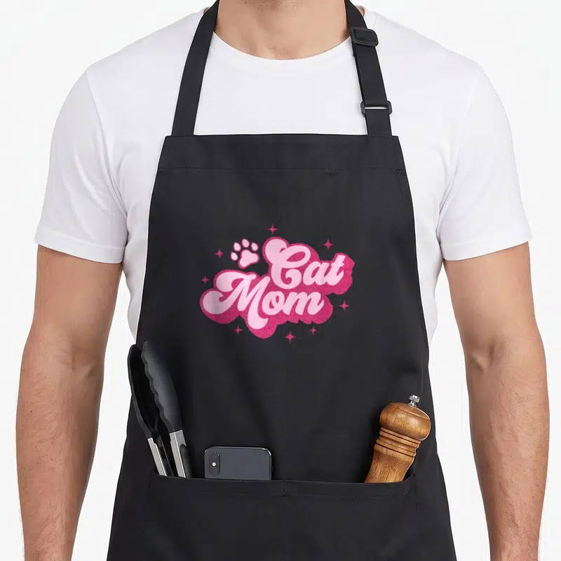 Mam kitty cat apron - Cat Mom text in light pink script on dark pink glittery shape with paw print and stars — shown on a black cotton canvas apron against a white background