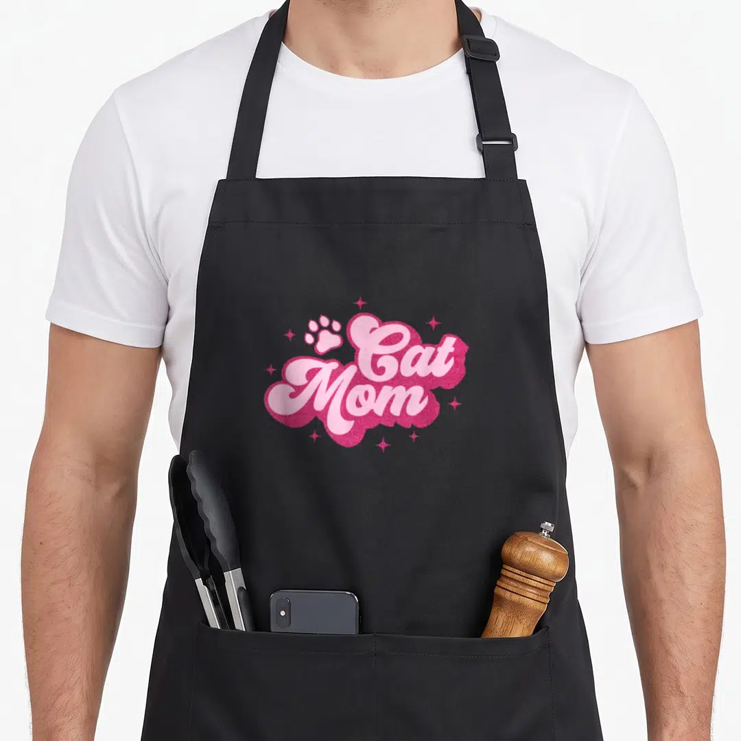 Mam kitty cat apron - Cat Mom text in light pink script on dark pink glittery shape with paw print and stars — shown on a black cotton canvas apron against a white background