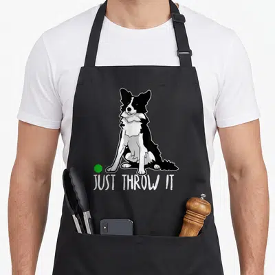 Throw border collie apron - Cartoon black and white dog sitting with green ball above JUST THROW IT text — shown on a black cotton canvas apron against a white background