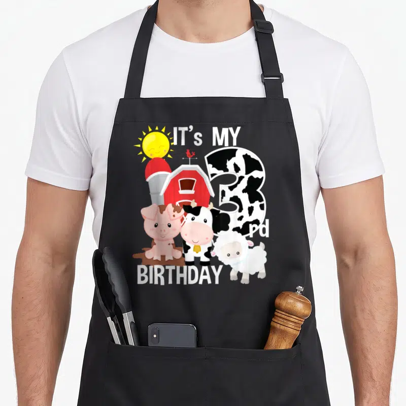 3rd farm aprons - Cartoon pig cow sheep barn cow print 3 birthday text — shown on a black cotton canvas apron against a white background