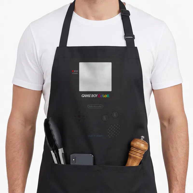 Comfort halloween apron - Front facing Game Boy Color console with grey body and blue select start text — shown on a black cotton canvas apron against a white background