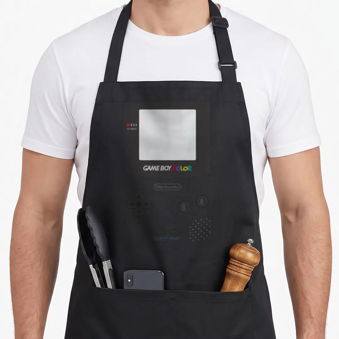 Comfort halloween apron - Front facing Game Boy Color console with grey body and blue select start text — shown on a black cotton canvas apron against a white background