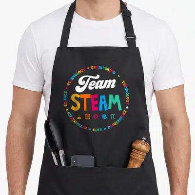 Steam school teacher apron - Team STEAM text with colorful circular border of subjects and five educational icons below — shown on a black cotton canvas apron against a white background