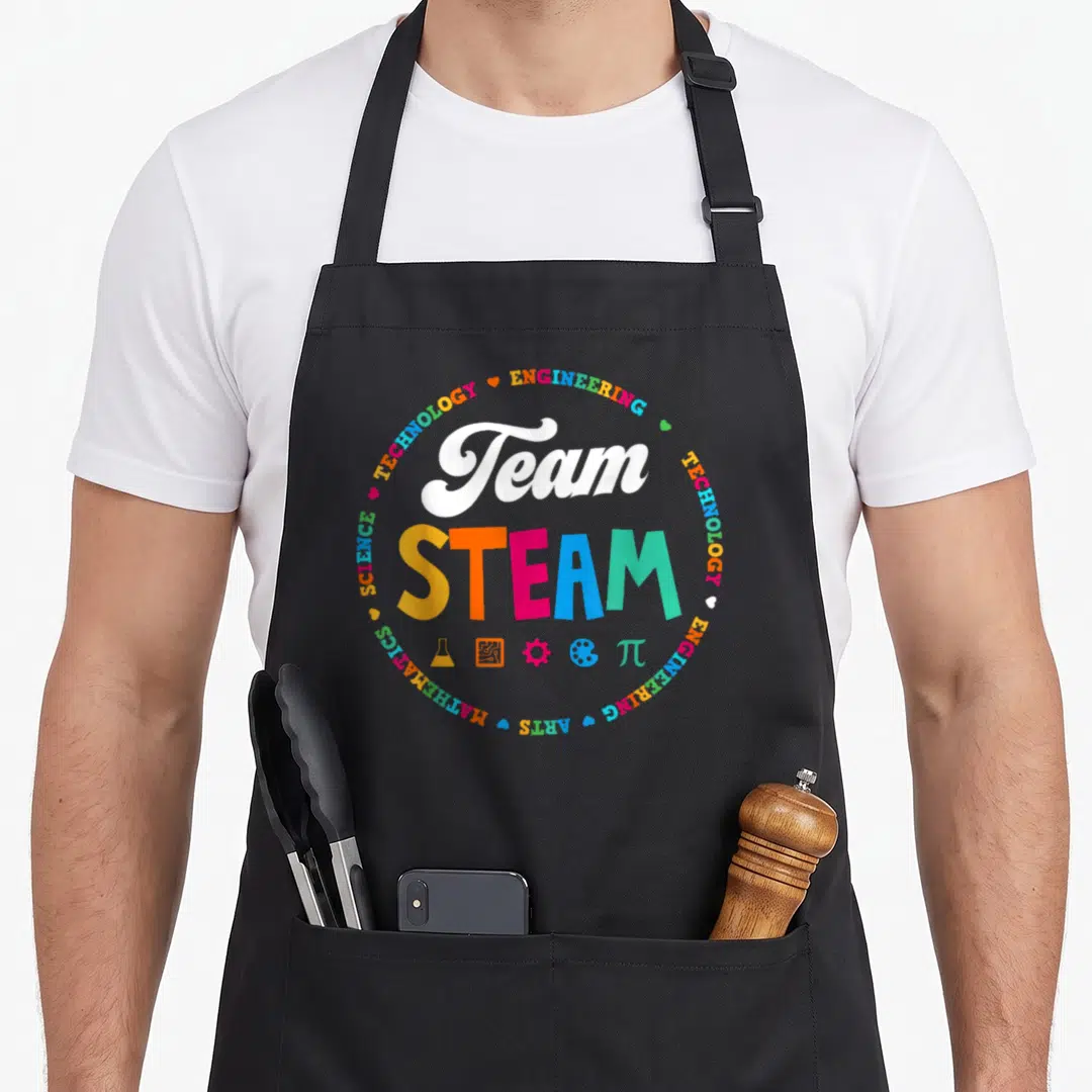 Steam school teacher apron - Team STEAM text with colorful circular border of subjects and five educational icons below — shown on a black cotton canvas apron against a white background