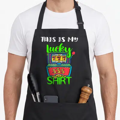 Slot casino aprons - Cartoon slot machine with 777 reels and text THIS IS MY Lucky SHIRT — shown on a black cotton canvas apron against a white background