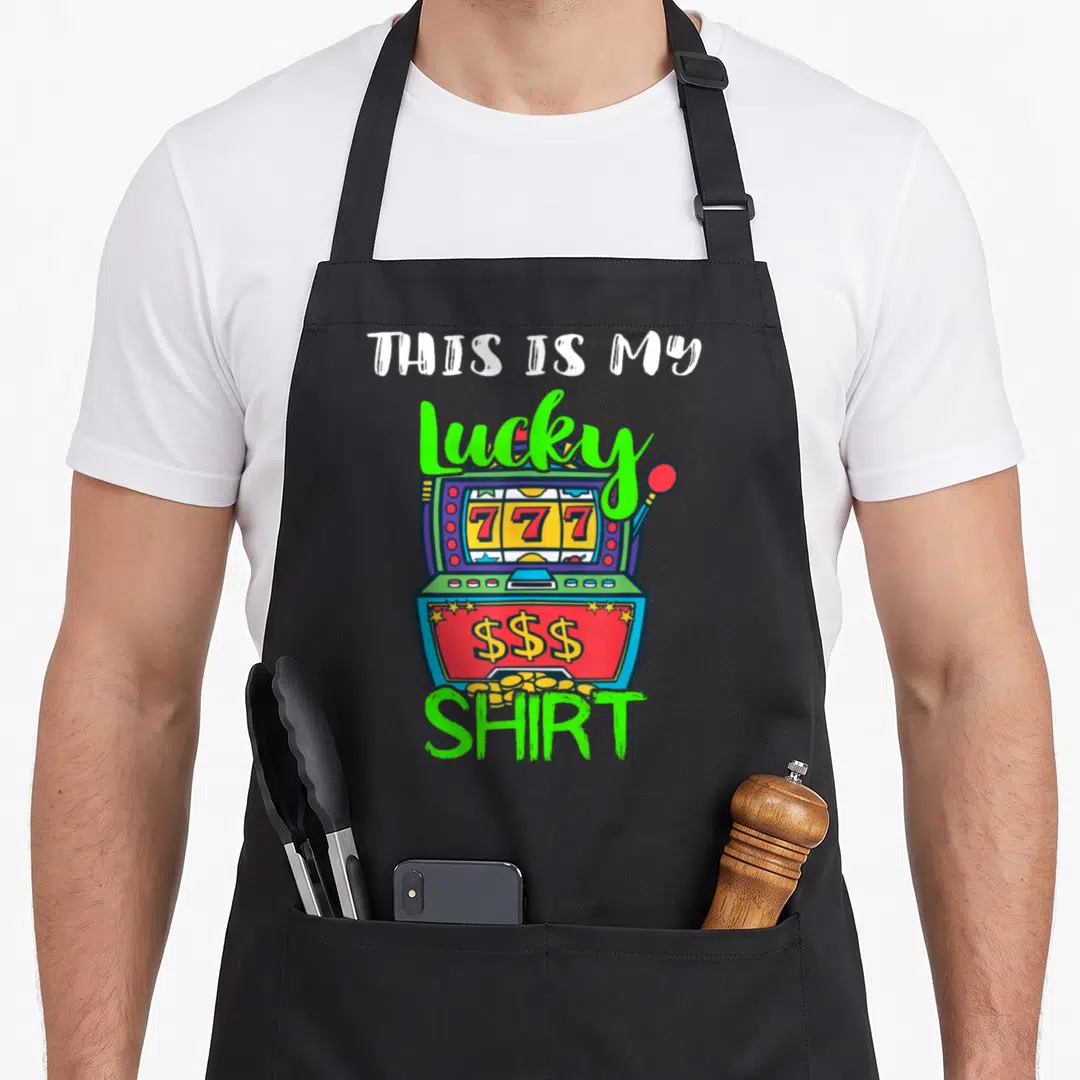 Slot casino aprons - Cartoon slot machine with 777 reels and text THIS IS MY Lucky SHIRT — shown on a black cotton canvas apron against a white background