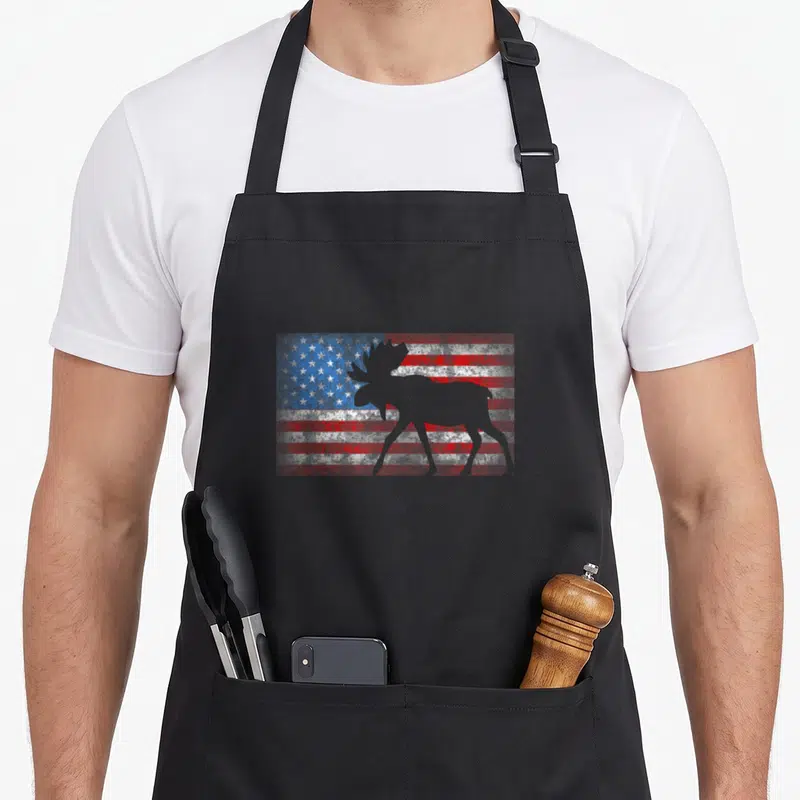 Moose 4th of july apron - Grey moose silhouette standing in front of a weathered red white and blue flag background — shown on a black cotton canvas apron against a white background