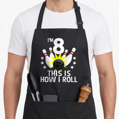Bowling birthday apron - I'M 8 text above black ball striking four pins with yellow burst and six stars — shown on a black cotton canvas apron against a white background