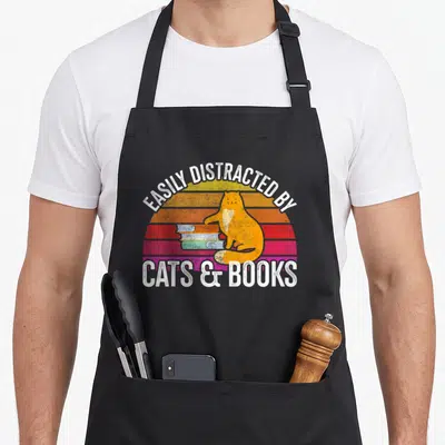 Avid cat apron - Orange cat sitting on books in front of striped sunset with retro text — shown on a black cotton canvas apron against a white background
