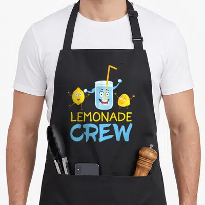 Boss lemonade apron - Cartoon glass of lemonade with arms flanked by two yellow lemons and stacked LEMONADE CREW text — shown on a black cotton canvas apron against a white background