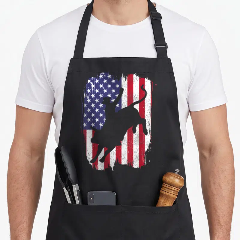 Bullriding american flag apron - Grey bull and rider silhouette over distressed red white blue American flag art — shown on a black cotton canvas apron against a white background