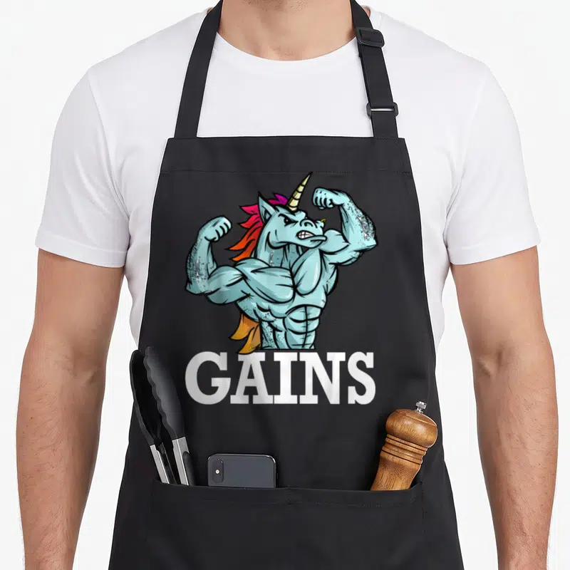 Gain unicorn apron - Muscular blue cartoon unicorn flexing arms above white GAINS text — shown on a black cotton canvas apron against a white background
