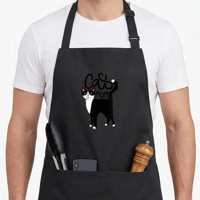 Cat animal tuxedo apron - Cartoon black and white cat with pink ears and curved Cat Mom text — shown on a black cotton canvas apron against a white background