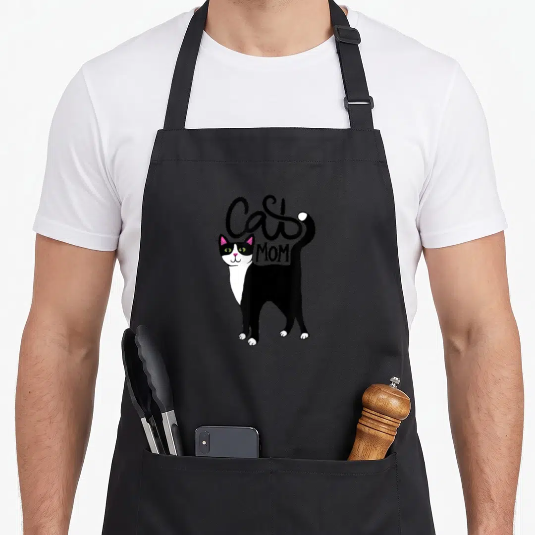 Cat animal tuxedo apron - Cartoon black and white cat with pink ears and curved Cat Mom text — shown on a black cotton canvas apron against a white background