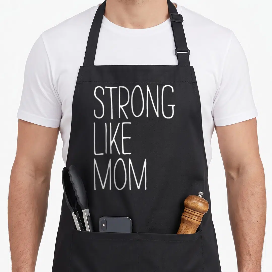 Funny strong mom apron - White stacked text reading STRONG LIKE MOM on grey background — shown on a black cotton canvas apron against a white background