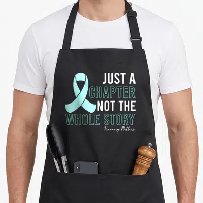 Manifest addiction recovery apron - Teal awareness ribbon and Just a Chapter Not The Whole Story text in white and teal — shown on a black cotton canvas apron against a white background