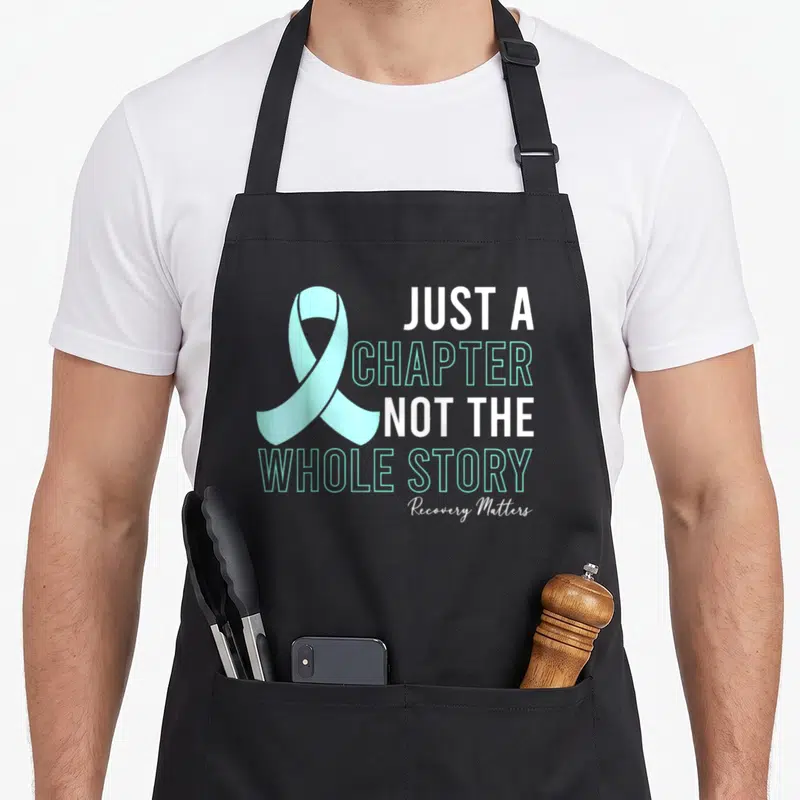 Manifest addiction recovery apron - Teal awareness ribbon and Just a Chapter Not The Whole Story text in white and teal — shown on a black cotton canvas apron against a white background
