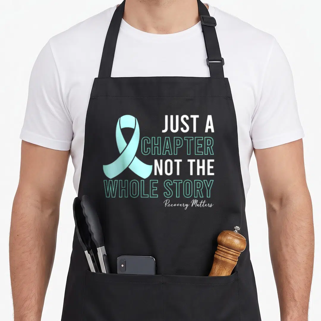 Manifest addiction recovery apron - Teal awareness ribbon and Just a Chapter Not The Whole Story text in white and teal — shown on a black cotton canvas apron against a white background
