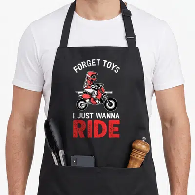 Toy bike apron - Cartoon motocross rider with arched text FORGET TOYS and stacked I JUST WANNA RIDE in red white black — shown on a black cotton canvas apron against a white background