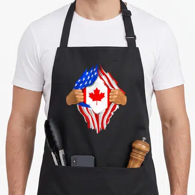 Canadian blood apron - Two brown hands tearing American flag to reveal red maple leaf Canadian flag — shown on a black cotton canvas apron against a white background