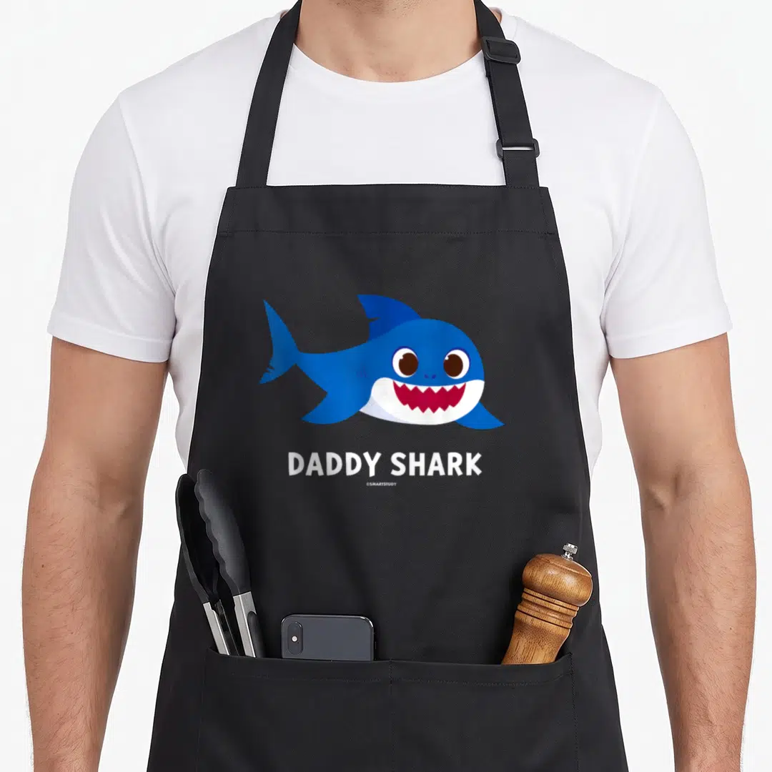 Pinkfong shark daddy apron - Blue cartoon shark illustration above white DADDY SHARK text — shown on a black cotton canvas apron against a white background